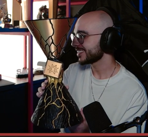 A smiling man wearing headphones and glasses proudly holds up an ornate, hourglass-shaped trophy with gold and black detailing. The image relates to "AURA," as the trophy's distinctive hourglass shape and the "Aura" branding on the microphone stand suggest he has just won an award or recognition for his content.
