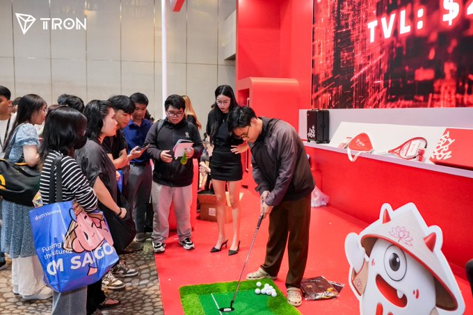 At the TRON-sponsored Conviction 2026 event in Ho Chi Minh City, a man putts golf balls on a miniature green while attendees gather around, with the TRON logo visible on the wall and a mascot displayed nearby.