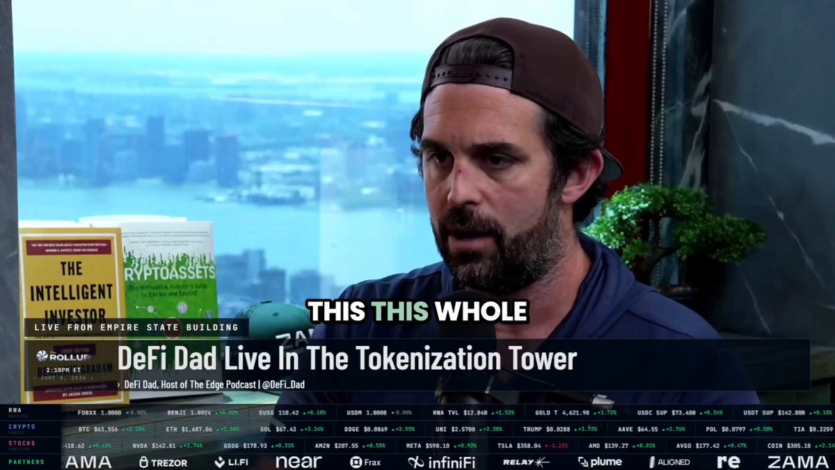 The image shows a man wearing a baseball cap, sitting at a desk with a TV screen behind him displaying an advertisement for Bitcoin. The text on the screen reads "This whole", which could be interpreted as a statement about the current state of DeFi or a reference to the Bitcoin advertisement.