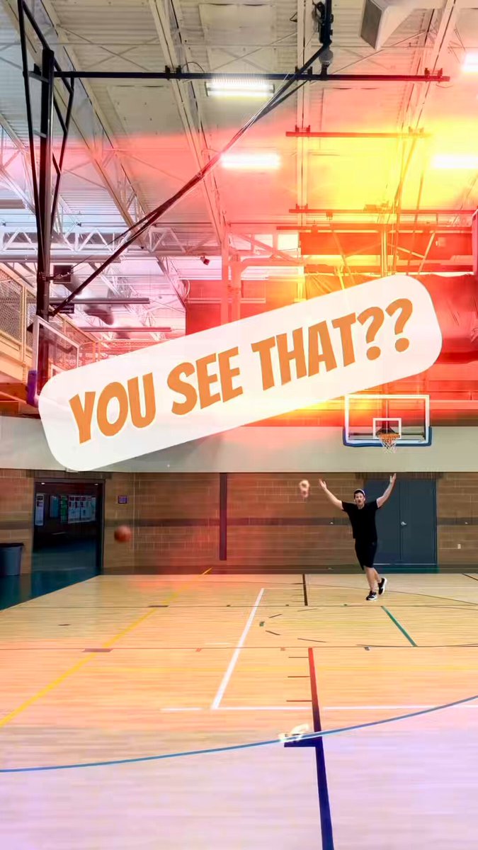 The image features a basketball court with a man standing near the basket, holding a basketball and preparing to shoot. The background of the photo includes a gymnasium with a basketball hoop mounted high above the ground. A banner can be seen hanging from the ceiling in the background, which reads "You see that?".