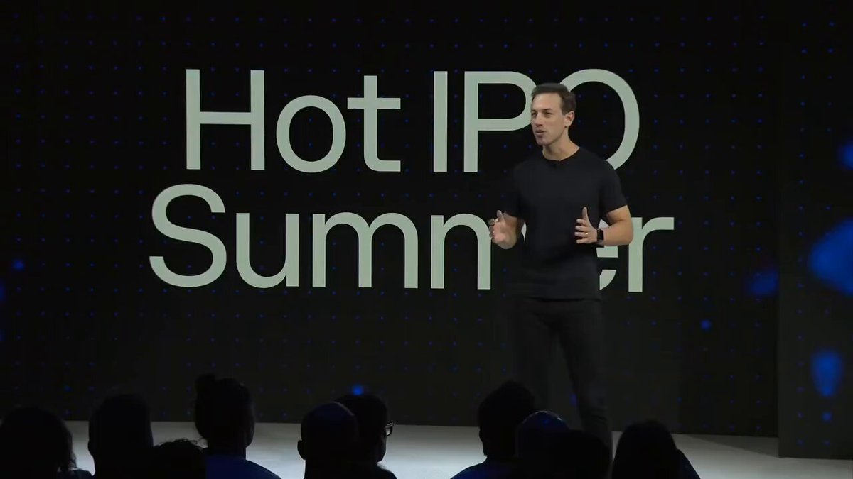The image shows a man standing on stage, giving a speech or presentation. He appears to be wearing a black shirt and pants. In front of him, there are several people sitting in chairs, possibly listening to his speech or watching a video related to the event. The background features a large screen displaying the text "Hot IPO SUMMER" along with some blue dots on it. This suggests that the image is related to an event where the man is speaking about cryptocurrency, specifically Bitcoin, and he might be discussing the IPO of Coinbase, as indicated by the post in the image.