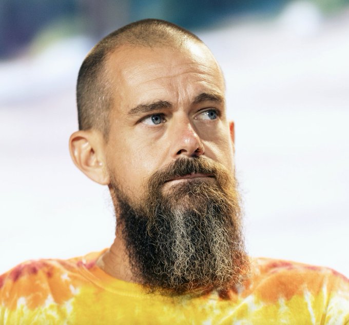 A close-up portrait of a man with a shaved head, long brown beard, and blue eyes, wearing a yellow and orange tie-dye shirt.
