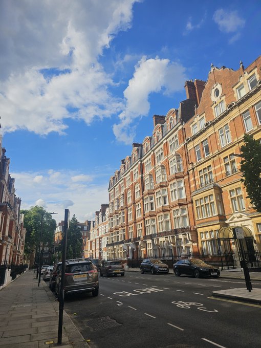 A sunny street scene on a London road with a 'BUS' lane, lined with elegant red brick Victorian-style buildings on the right and parked cars along the curb.