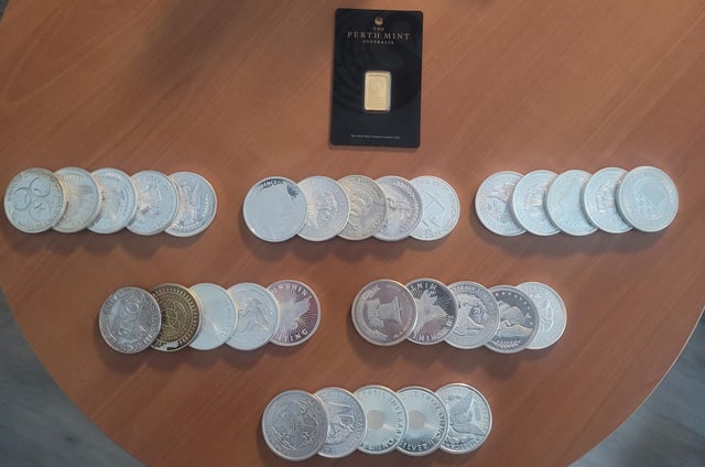 A collection of silver coins and a 1g Perth Mint gold bar are arranged on a wooden table, representing a starter stack of precious metals. The display showcases various silver pieces—including what appear to be Britannias, maples, and other bullion designs—alongside the small gold bar from Perth Mint, marking the beginning of a precious metals stacking journey.