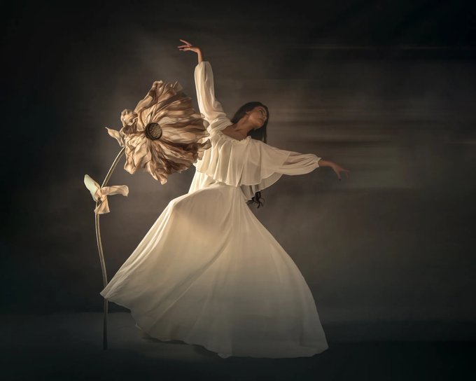 A dancer in a flowing white dress poses gracefully beside a large, golden artificial flower against a dark, moody background with motion blur effects.