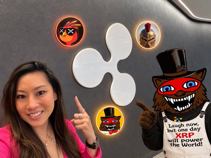 A smiling woman in a pink top points up at a Ripple (XRP) logo displayed on a wall, with the caption "Laugh now, but one day XRP will power the World!" visible on a cartoon cat figure beside her. The image visually supports the post's narrative by showing the woman gesturing directly at the Ripple logo, suggesting her engagement with XRP.