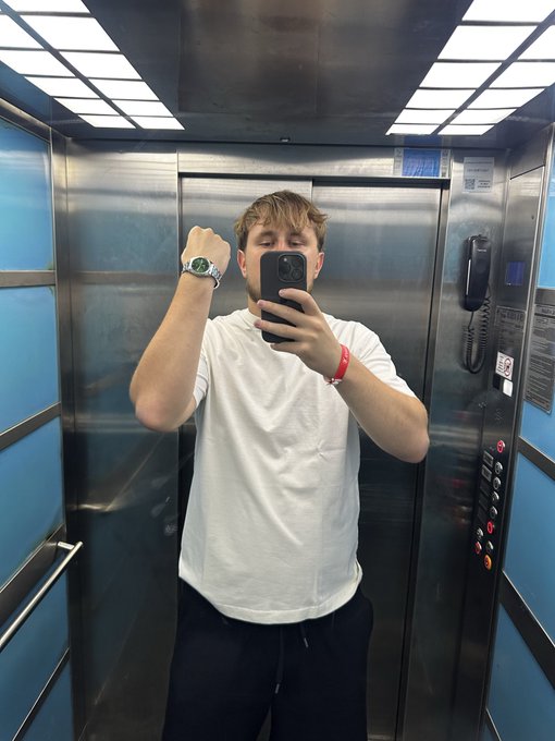 A young man in a white t-shirt takes a mirror selfie with his phone inside an elevator, showing off a watch on his raised wrist.