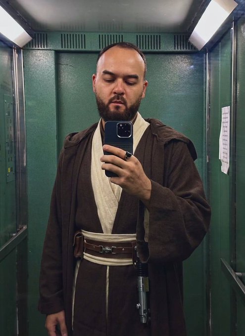 A bearded man in a Jedi-style brown robe takes a mirror selfie with his iPhone in an elevator, appearing to embody the Star Wars character in a humorous cosplay. The image ties into the post's playful theme of waiting for $CHOG to reach $1, with the Jedi pose seemingly representing a hopeful, patient stance like watching over the moon-bound token.