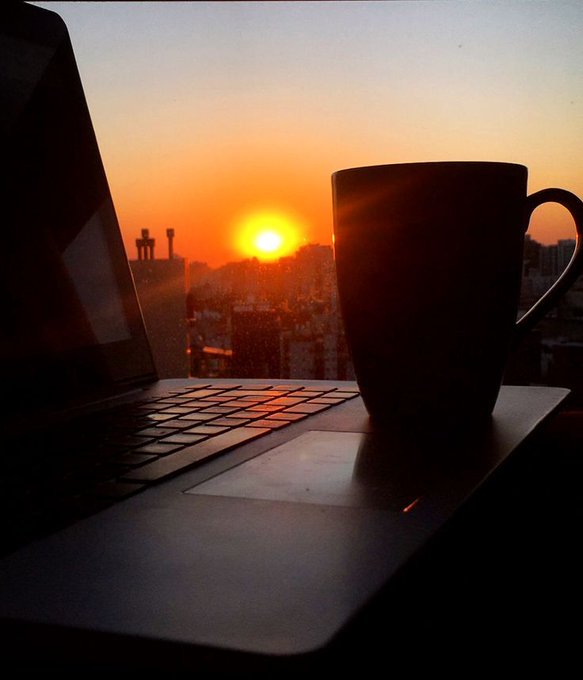 A silhouette of a coffee mug and an open laptop sits on a windowsill against a vibrant orange sunset over a cityscape.