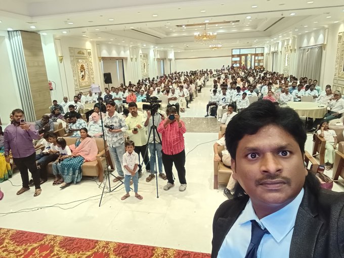 A man in a suit takes a selfie in the foreground at a packed indoor hall, where a large crowd is seated facing a stage area, with several people including a child standing near a tripod-mounted camera. The image captures the energetic community gathering referenced in the post, showing the full venue filled with attendees celebrating the 10 Million Linkers milestone in India.