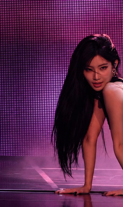 A woman with long dark hair kneels on a stage under pink lighting, looking intensely at the camera.