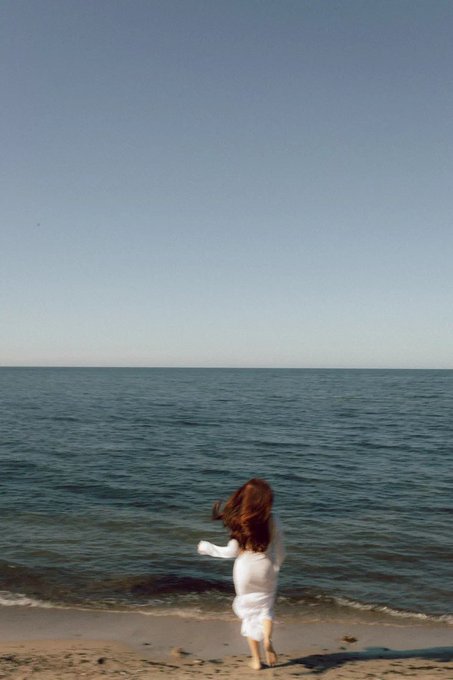 A vintage-style photograph shows a young child with long brown hair running barefoot along a sandy beach toward the calm ocean waters under a clear blue sky.