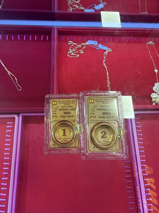 The image shows a display case filled with gold jewelry items such as necklaces and bracelets. There are two gold bracelets prominently displayed inside clear plastic cases, each labeled with numbers 1 and 2. The background of the image features a pinkish-red color scheme, which complements the gold jewelry on display.