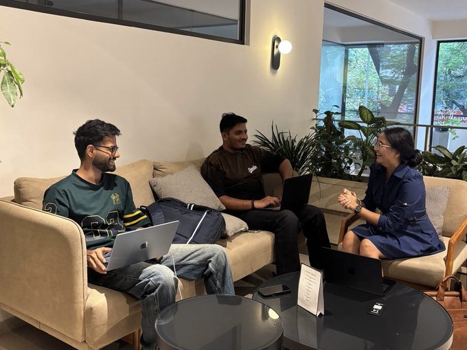 Three people are seated on couches in a modern lounge area with plants, engaged in a conversation; one person is using a laptop.
