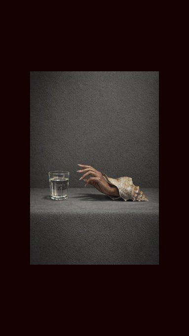 A surreal image shows a hand wearing a beaded sleeve reaching toward a glass of water on a gray surface against a dark background.