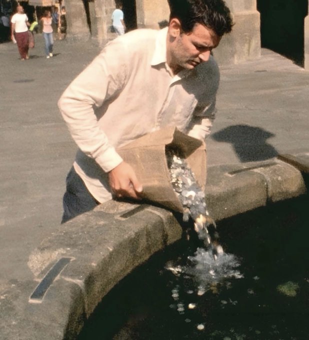 A man pouring a large sack of coins into a stone fountain or trough, with two people walking in the background.