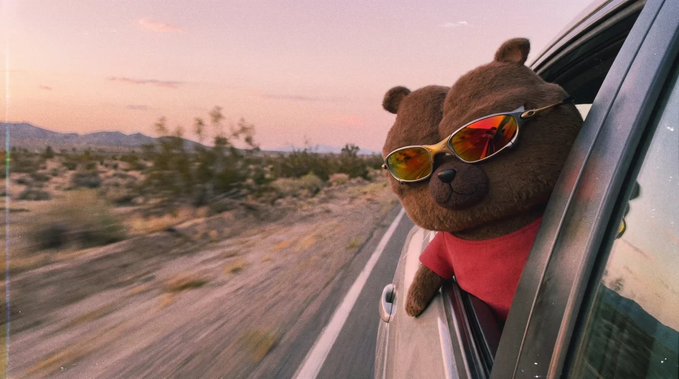 A teddy bear wearing sunglasses and a red shirt leans out of a car window along a desert road at sunset.