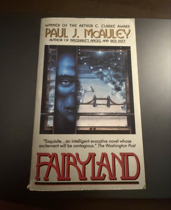 A paperback book titled 'FAIRYLAND' by Paul J. McAuley, winner of the Arthur C. Clarke Award, featuring cover art of a face looking out at a bridge and cityscape.