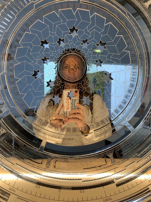 A creative photograph showing a person taking a picture with a smartphone, framed within the circular face of a watch or dial featuring a central Bitcoin logo surrounded by star cutouts.
