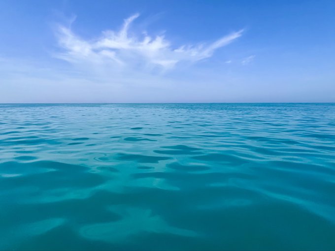 A serene view of turquoise ocean water stretches to the horizon under a bright blue sky with wispy white clouds. Gentle ripples reflect on the calm sea surface.