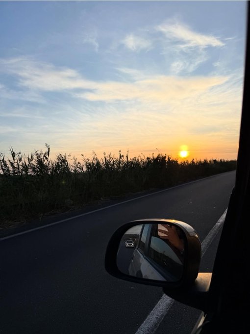 A scenic sunset view captured from inside a moving vehicle, with tall grass lining the road and a side mirror reflecting the person taking the photo.