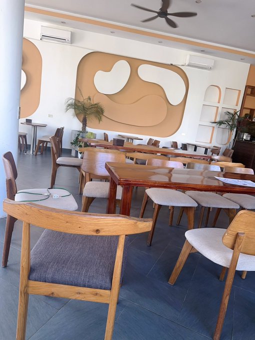 An empty restaurant interior features wooden tables and chairs arranged on tiled flooring, with distinctive tan-colored artistic wall designs and a ceiling fan overhead.
