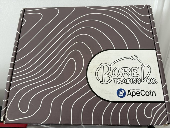 A brown cardboard box with white topographic line patterns features a label reading "BORED TRADING CO." powered by ApeCoin, indicating a branded shipping package.