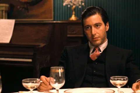 A man in a dark suit and orange tie sits at an elegant dinner table with wine glasses and a piano in the background, appearing serious and contemplative. The image is likely used as a stylistic, cinematic visual to lend a sophisticated, "insider" tone to the post promoting NFT communities and creators—mimicking a dramatic, mob-boss-like aesthetic to suggest authoritative or exclusive crypto advice.