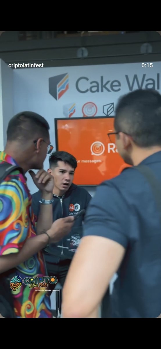 Several people are gathered at a Cake Wallet booth during the Crypto Latin Fest event, with a young man in a branded hoodie speaking to attendees near a screen displaying the wallet's interface.