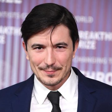 Portrait of a man with dark hair and a mustache wearing a dark suit, white shirt, and dark tie, posed in front of a step-and-repeat backdrop.