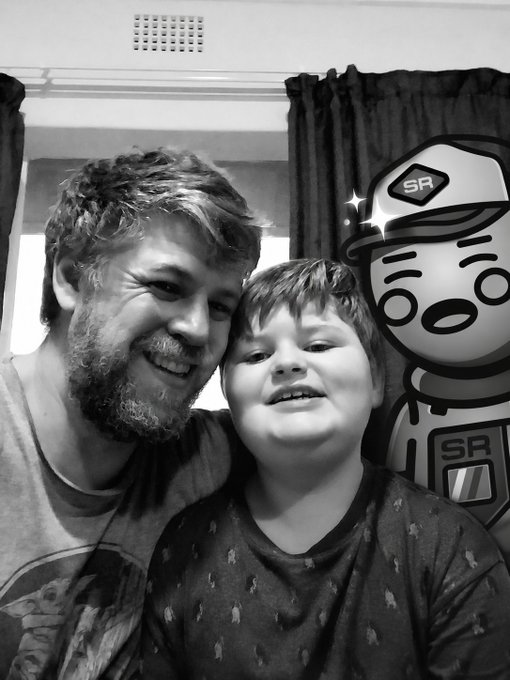 A black and white selfie of a smiling bearded man and a young boy, accompanied by a cartoon character wearing a cap with an 'SR' logo.