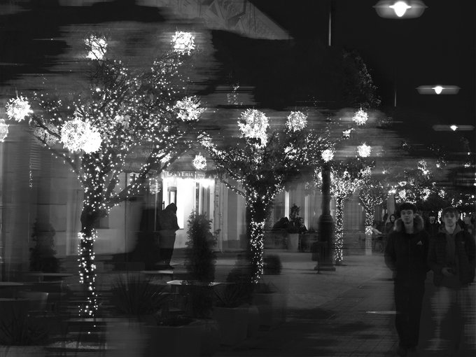A black and white photograph of a street scene at night, featuring trees decorated with sparkling lights, hanging star lanterns, and people walking by.