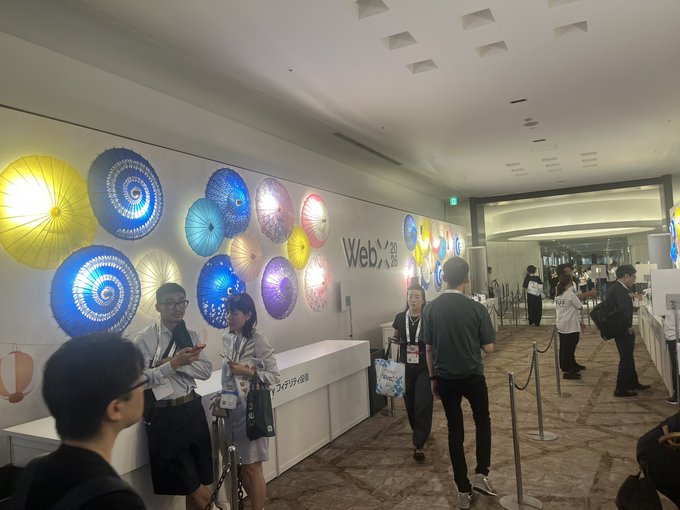 The image shows the registration or entrance area of "WebX" (with Japanese text "ウェブエックス" below it), featuring colorful illuminated umbrella-like disc decorations on the wall in blue, yellow, and other hues, with attendees milling about near stanchion barriers. The caption about Japan's WebX atmosphere fits well, as the scene captures that pre-event buzz—with crowds gathering and networking in a polished venue while conveying a sense that the institutional presence may be more performative than action-oriented.