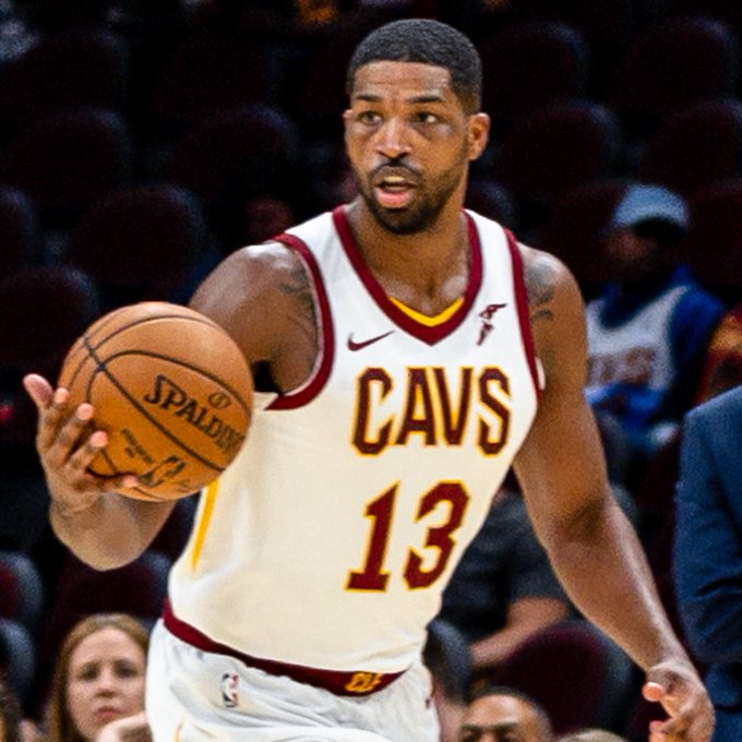 The image features a basketball player wearing a white and red uniform, holding a basketball while standing on a court. The player's jersey number is 13. In the background, there are several people watching the game or waiting for their turn to play. A person in the crowd has a name tag that reads "Tristan Thompson".