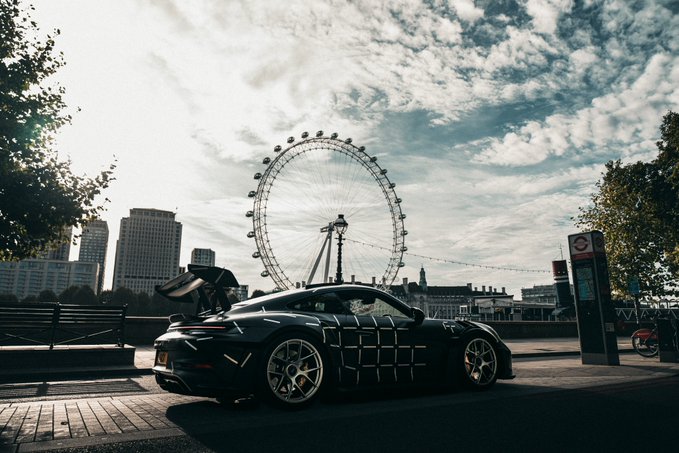 A sleek black Porsche 911 GT3 RS with its rear spoiler raised is parked along the Thames, with the London Eye looming in the background against a partly cloudy sky. The image captures the sports car the post is offering rides in around London on 14 November to those who purchase a Solana Breakpoint ticket with code BP26-MATTHEW.