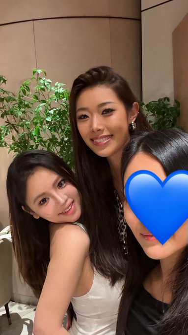 Three women pose closely together for a selfie, with green plants and beige walls in the background; one person's face is obscured by a blue heart emoji, matching the post's shoutout to ShirleyXBT and XtineFang from Polymarket.