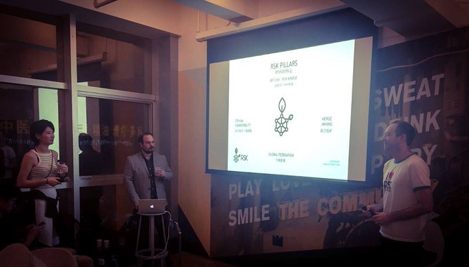 A photo from a 2017 meetup in China where a presentation on "Risk Pillars" and "Smarter Bitcoin" (RSK) is being given, with two men presenting beside a projected slide about Bitcoin staking, community integrity, and hedge mining, contextualized by the post as an early example of global Bitcoin programmability efforts before mainnet.