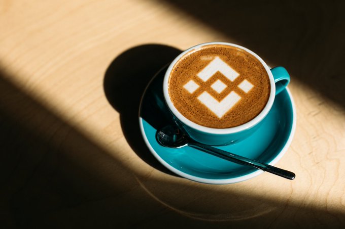 A turquoise coffee cup with latte art featuring the Binance logo (a stylized 'B' made of diamond shapes) sits on a wooden table in sunlight.