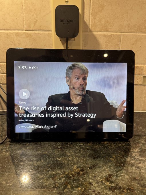 An Amazon Echo Show displays a Yahoo Finance news video titled "The rise of digital asset treasuries inspired by Strategy," featuring Michael Saylor, with a prompt reading "Try 'Alexa, what's the story?'"—suggesting the device is promoting Bitcoin/crypto content. The image humorously illustrates how Alexa is "orange pilling" users by surfacing pro-Bitcoin news coverage.