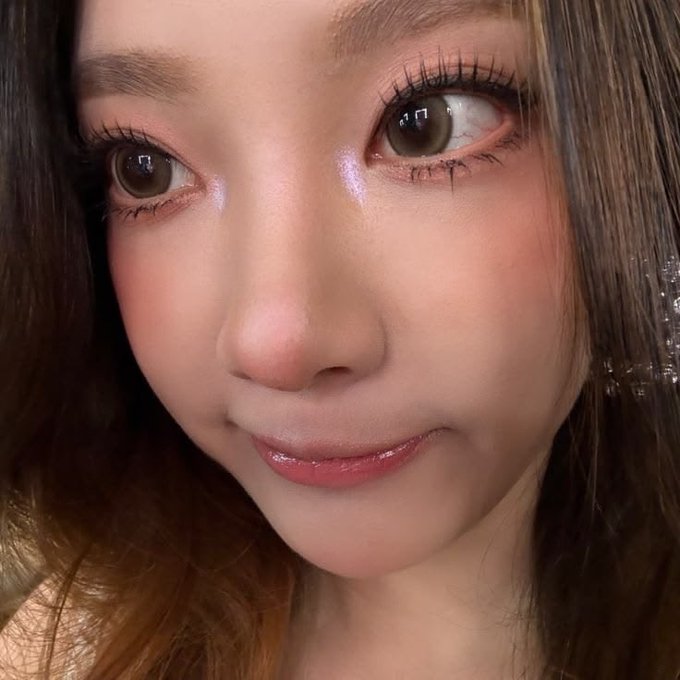 A close-up selfie of a person with brown eyes, rosy cheeks, glossy pink lips, and long eyelashes.