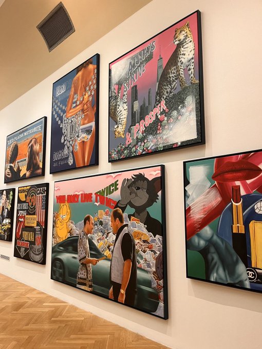 A gallery wall display featuring various framed pop art and contemporary artworks, including pieces with leopards, dollar bills, and figures, hung above a light wood herringbone floor.