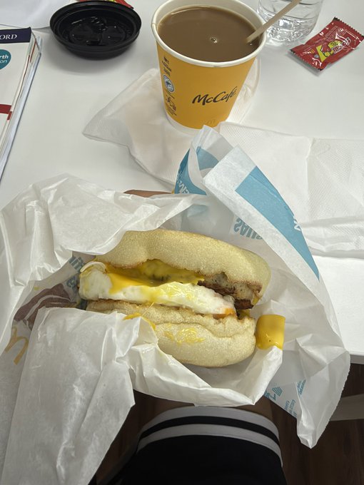 A McCafé coffee in a yellow cup sits next to an unwrapped breakfast sandwich with egg, cheese, and sausage patty on a paper wrapper.