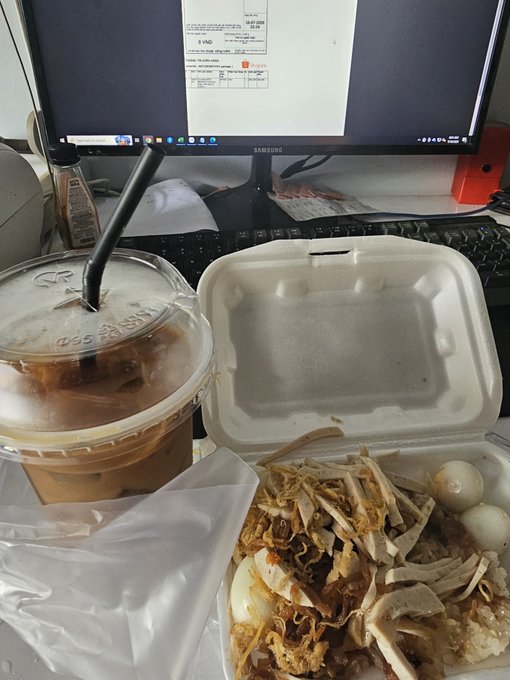 A desk scene showing an iced coffee in a plastic cup, an open takeout container with rice, chicken, egg, and quail eggs, with a Samsung monitor displaying a FedEx form in the background.