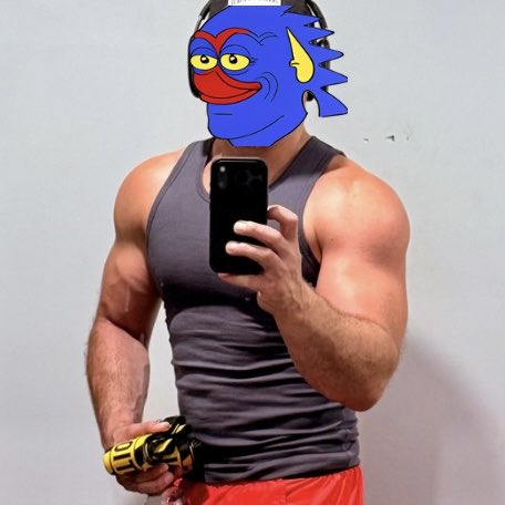A muscular man takes a mirror selfie wearing a gray tank top, red shorts, and a cartoon fish mask over his face, holding a phone and a yellow item.