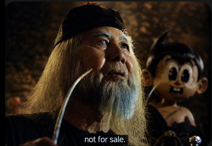 A cinematic still showing an older man with long white hair and beard wearing a black cap, with a small dark doll-like figure beside him; subtitle reads 'not for sale.'