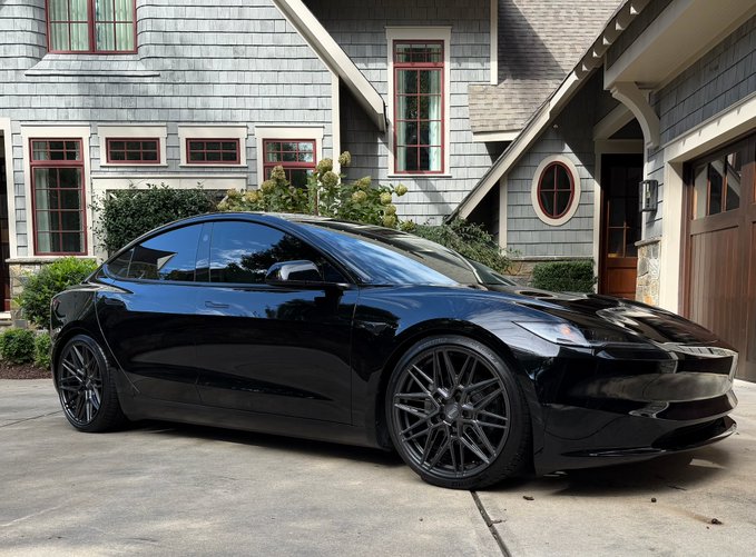 A black Tesla sedan parked in a driveway in front of a gray-shingled house with red-trimmed windows.