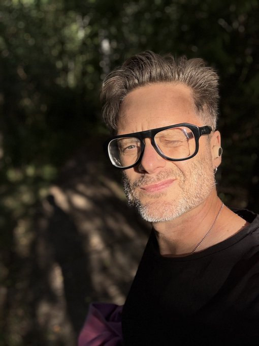 A portrait of a man with salt-and-pepper hair and beard, wearing black glasses and a black shirt, smiling outdoors in warm sunlight.