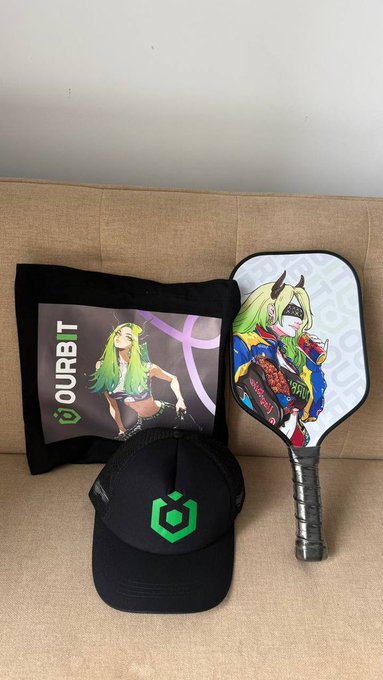 Branded merchandise from the crypto project 'OURBIT' displayed on a couch, including a pillow, a cap, and a pickleball paddle featuring anime-style artwork.
