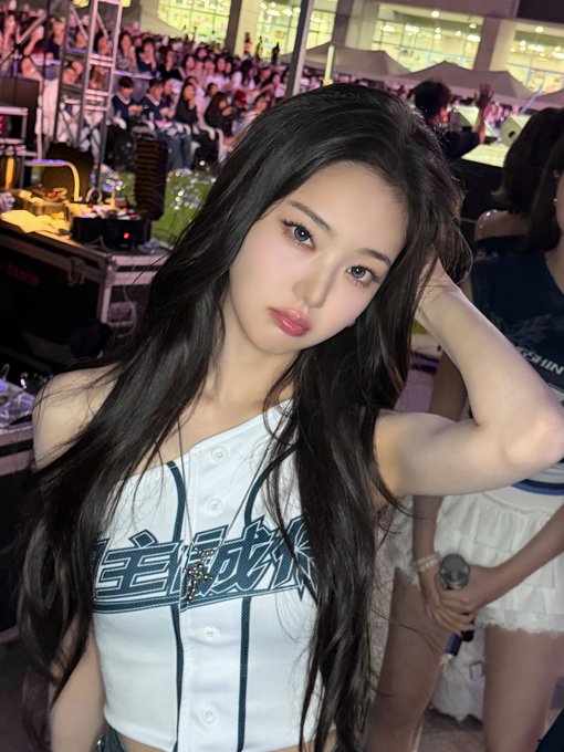 A woman with long dark hair poses for a selfie at a concert venue, wearing a white and navy baseball-style crop top with Asian characters on the front.