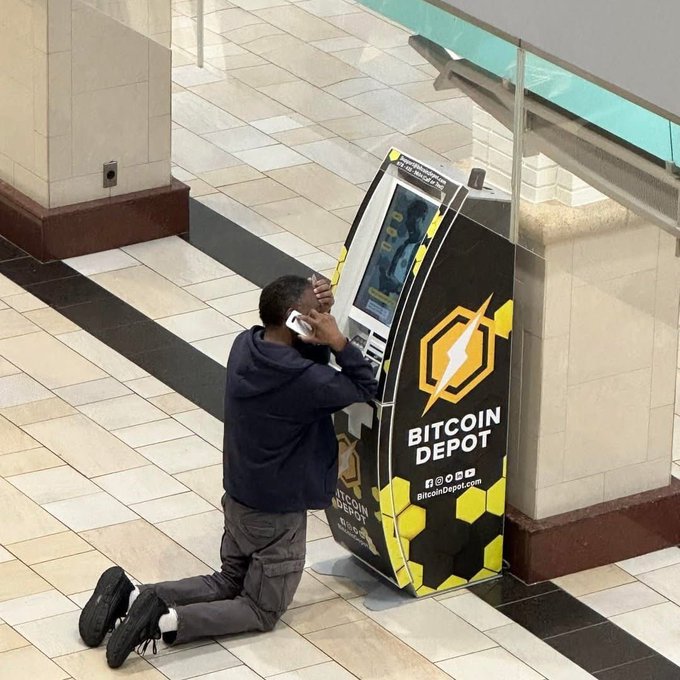 A man kneels on the floor of a mall next to a Bitcoin Depot ATM, appearing distressed while holding a phone to his ear with one hand and his head with the other.